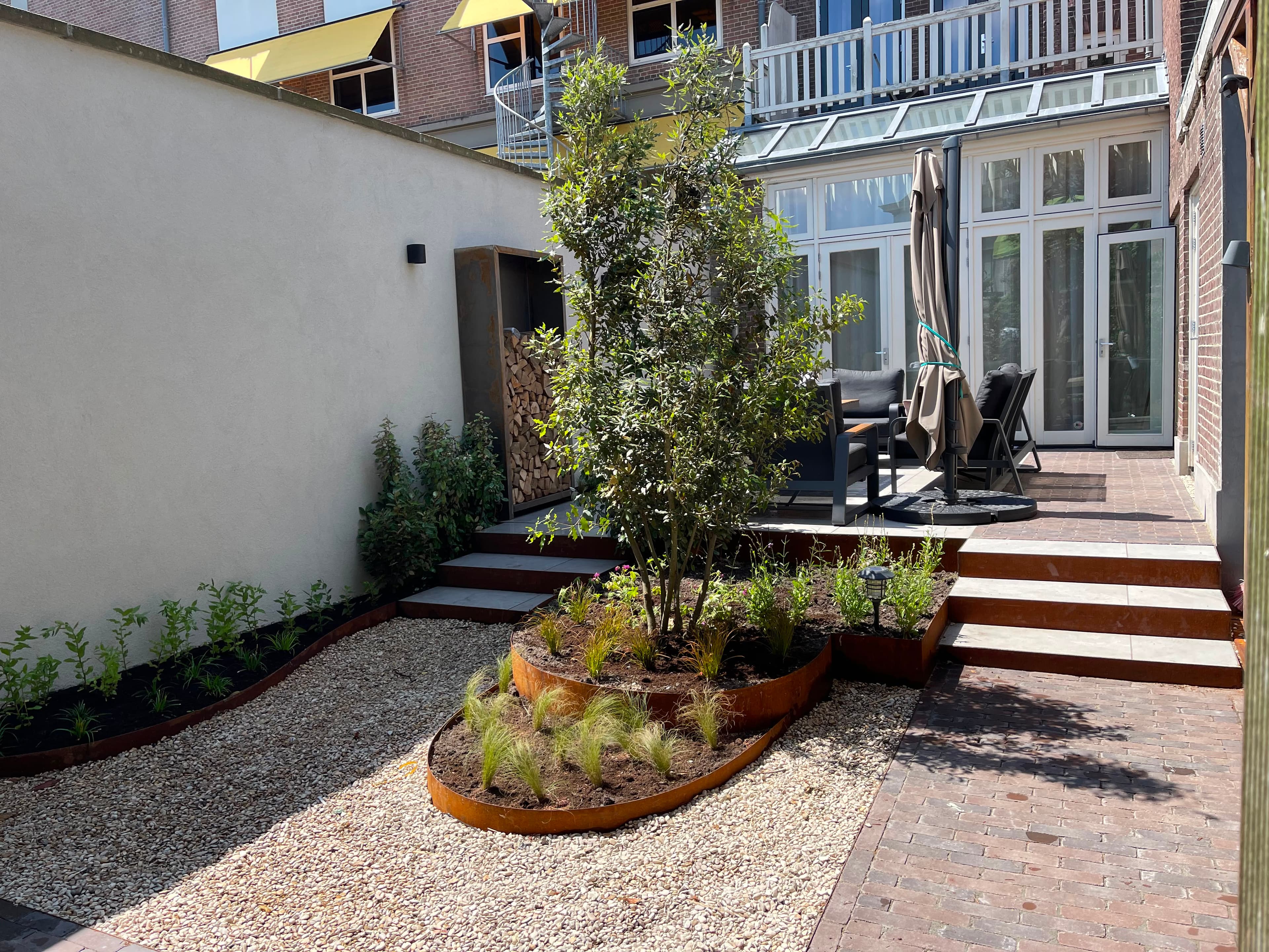 Zonnige tuin van Steengoed Tuinen met cortenstalen plantenbakken, olijfboom en grind